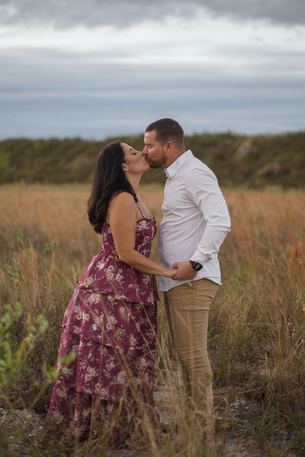 A romantic couple embraces in tall grass during an overcast day, with her wearing a floral mauve dress and him in white.