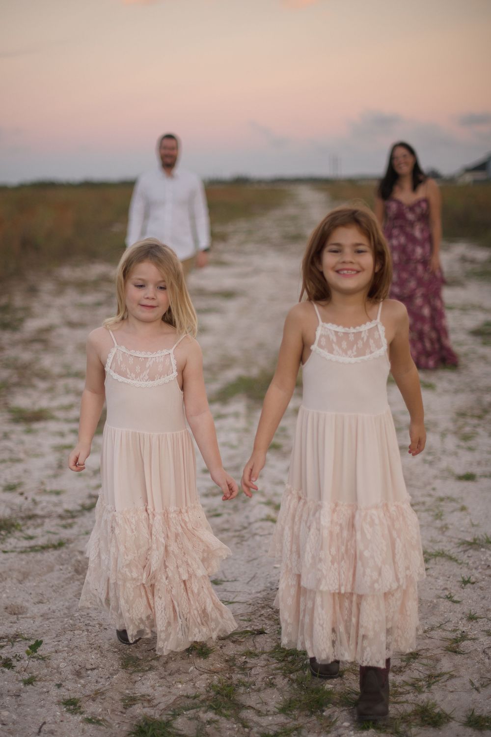 Children holding hands walk along a sandy beach path at sunset wearing matching lace dresses in soft pink tones.