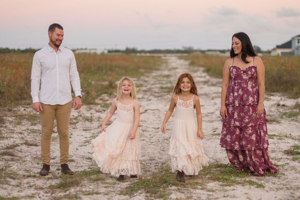 A family of four poses together in coordinating outfits on a sandy beach path at sunset.