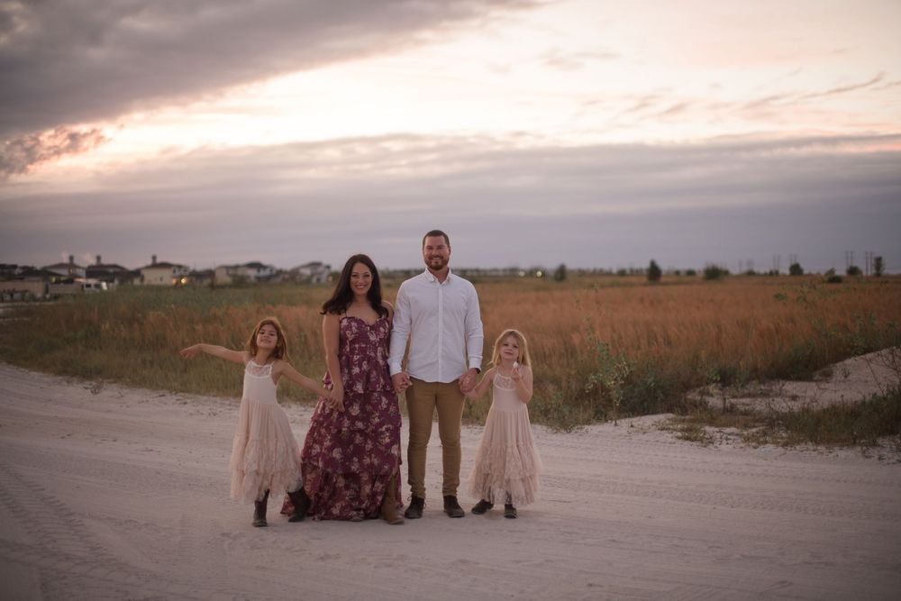 A family of four poses together on a dirt road at sunset with tall grass and suburban houses in the background.