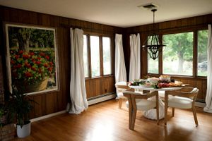 A cozy dining room with wood paneling, white curtains, and a colorful floral painting on the wall.