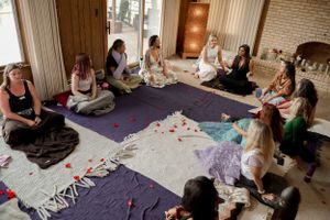Group seated in circle on purple blankets during an indoor gathering.