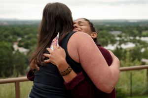Two people sharing an emotional hug on an outdoor observation deck overlooking a scenic landscape.