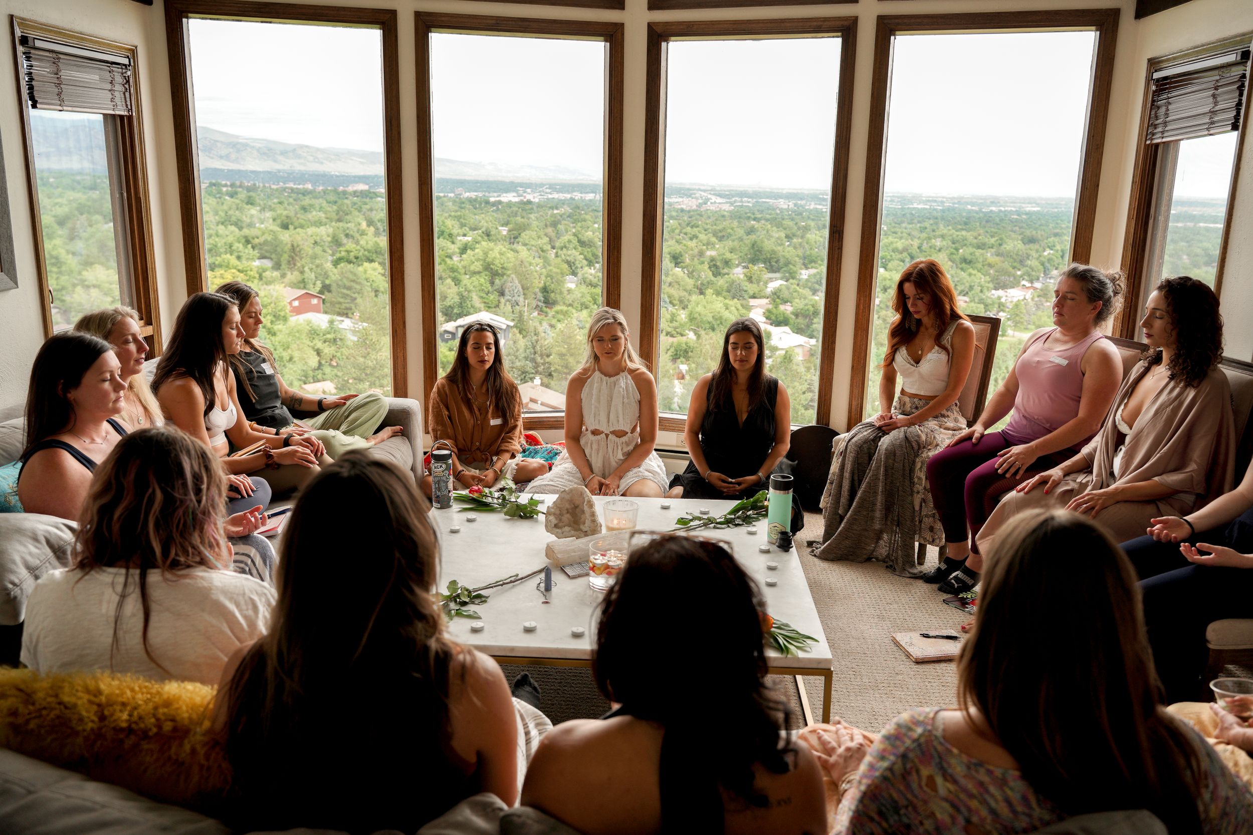 Large group seated in circular formation in bright room with floor-to-ceiling windows.