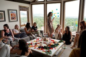A group gathers in a bright room with large windows for a casual social gathering with food and drinks.