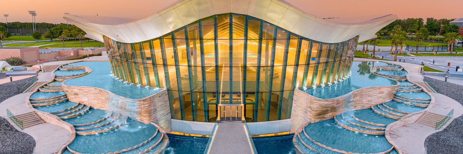 Aerial panorama of a luxurious aquatic center with winding pools, water features and modern glass buildings at sunset.