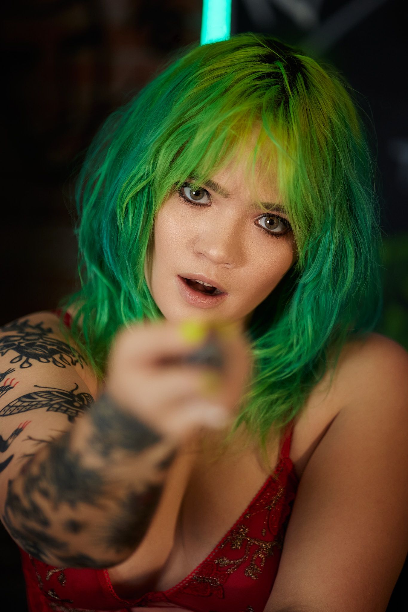 Woman with bright green hair wearing a red dress has visible tattoos on her arm while pointing at the camera.