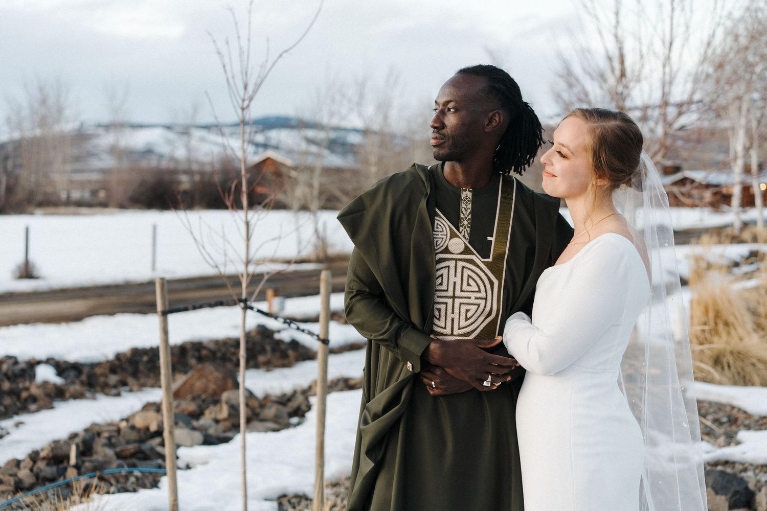 A couple in wedding attire standing together in a snowy winter landscape with rustic wooden fencing in the background.