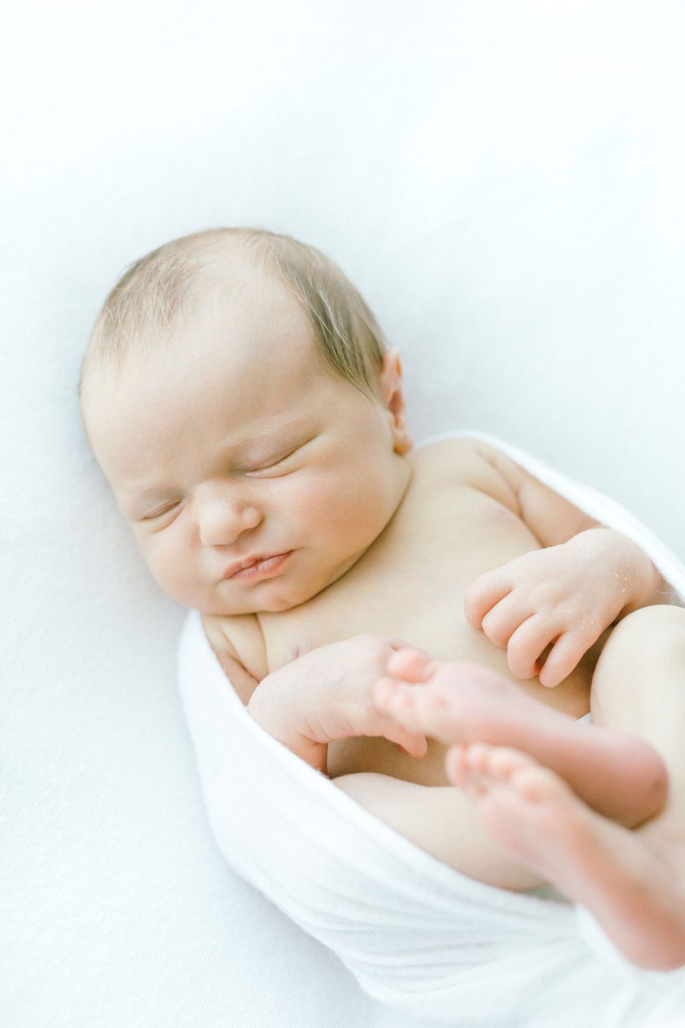 Sleeping newborn baby wrapped in white fabric with soft natural lighting.
