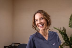 Healthcare worker in navy scrubs smiles warmly against neutral background with decorative plant.