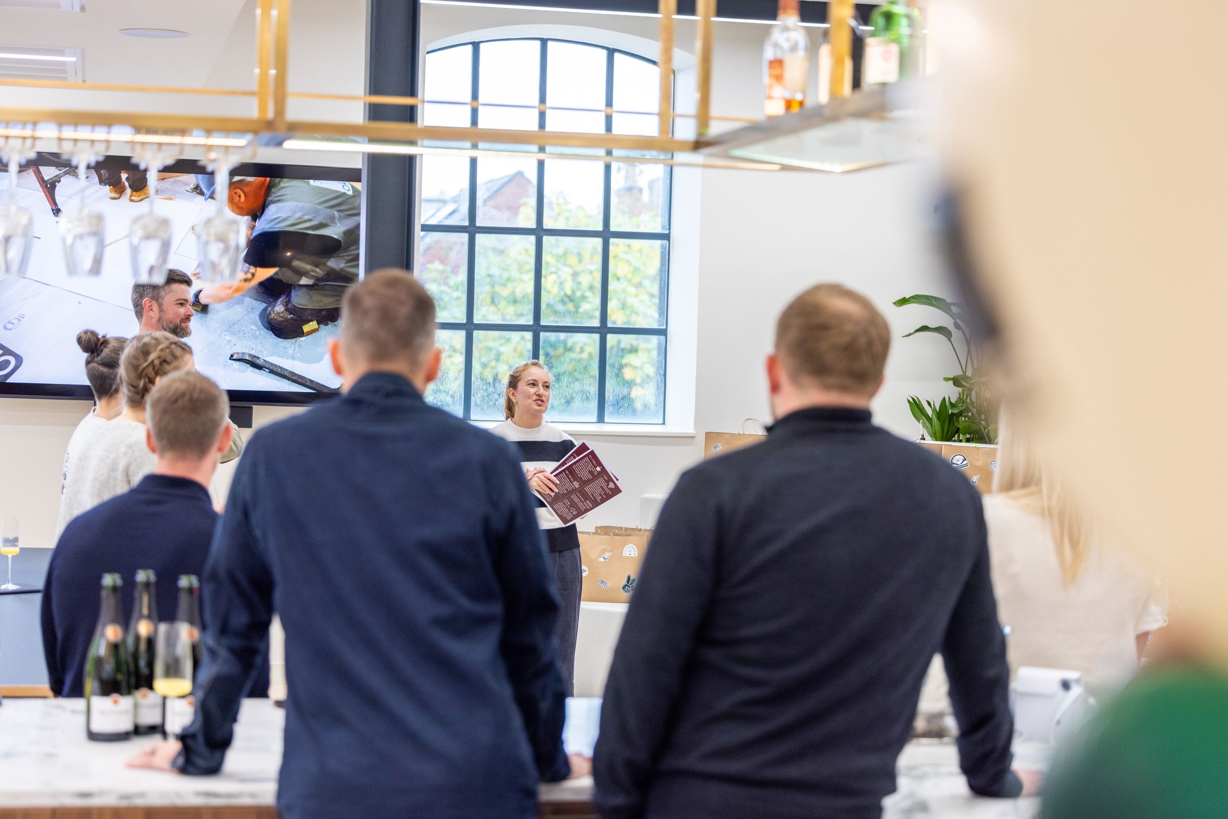 People gathered at a bar counter viewing menus while wine glasses hang overhead in a bright modern space.