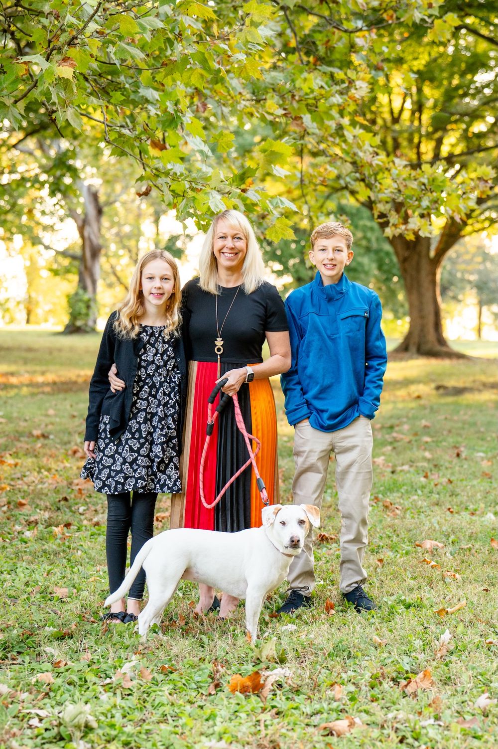 Group portrait with white dog surrounded by autumn leaves in a park setting on a sunny afternoon.