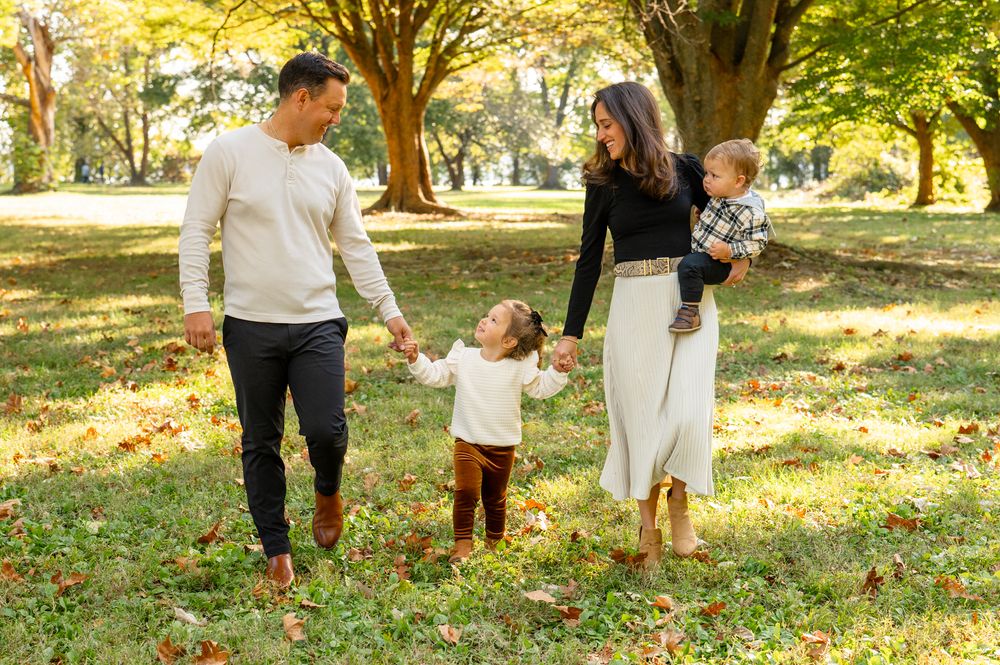 A family walks together holding hands on fallen autumn leaves in a sunlit park on a beautiful fall day.