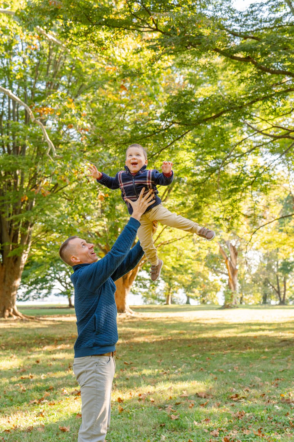 A joyful moment captured in a sunlit park with autumn trees as dad lifts his son up high in the golden light.