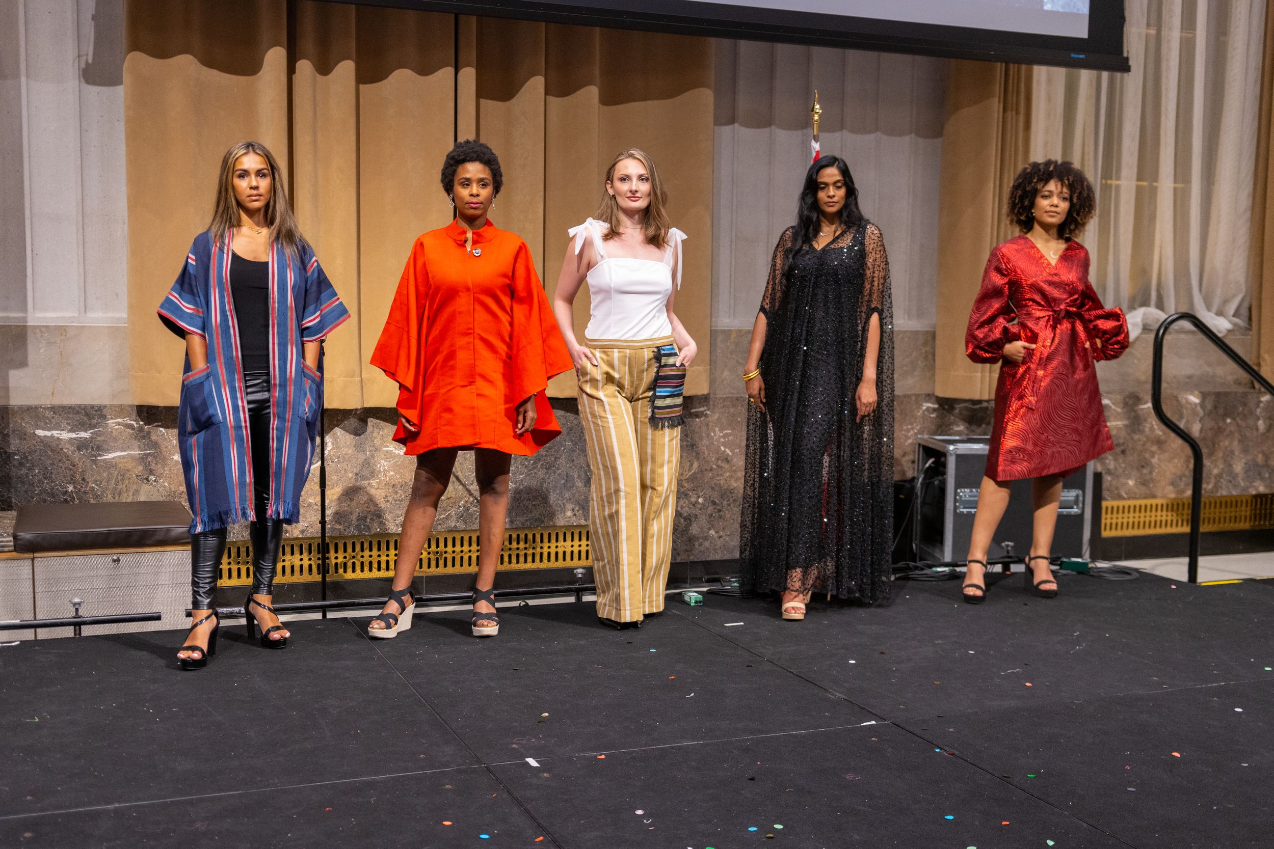 Models showcase diverse fashion styles including denim, orange dress, white top with khaki pants, black maxi dress and red dress on stage.