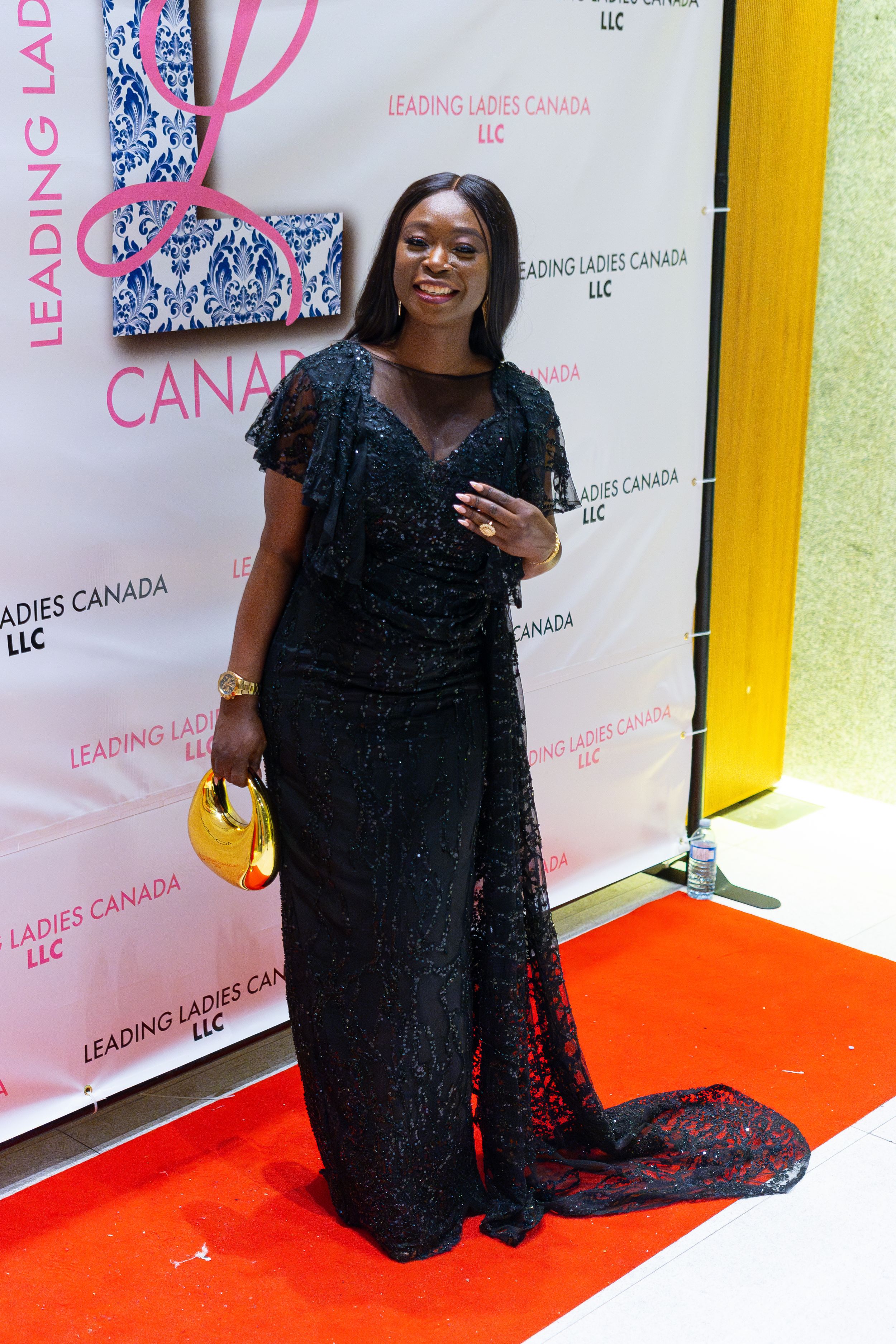 Elegant guest poses on red carpet wearing sparkling black evening gown with train and holding yellow clutch at Leading Ladies event.