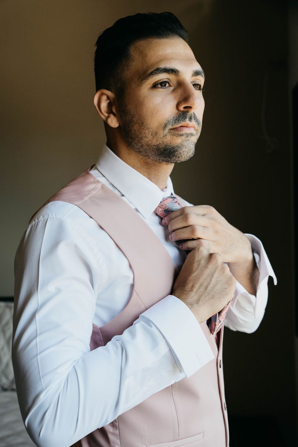 A person wearing a crisp white dress shirt and pink vest adjusts their tie in a formal portrait setting.
