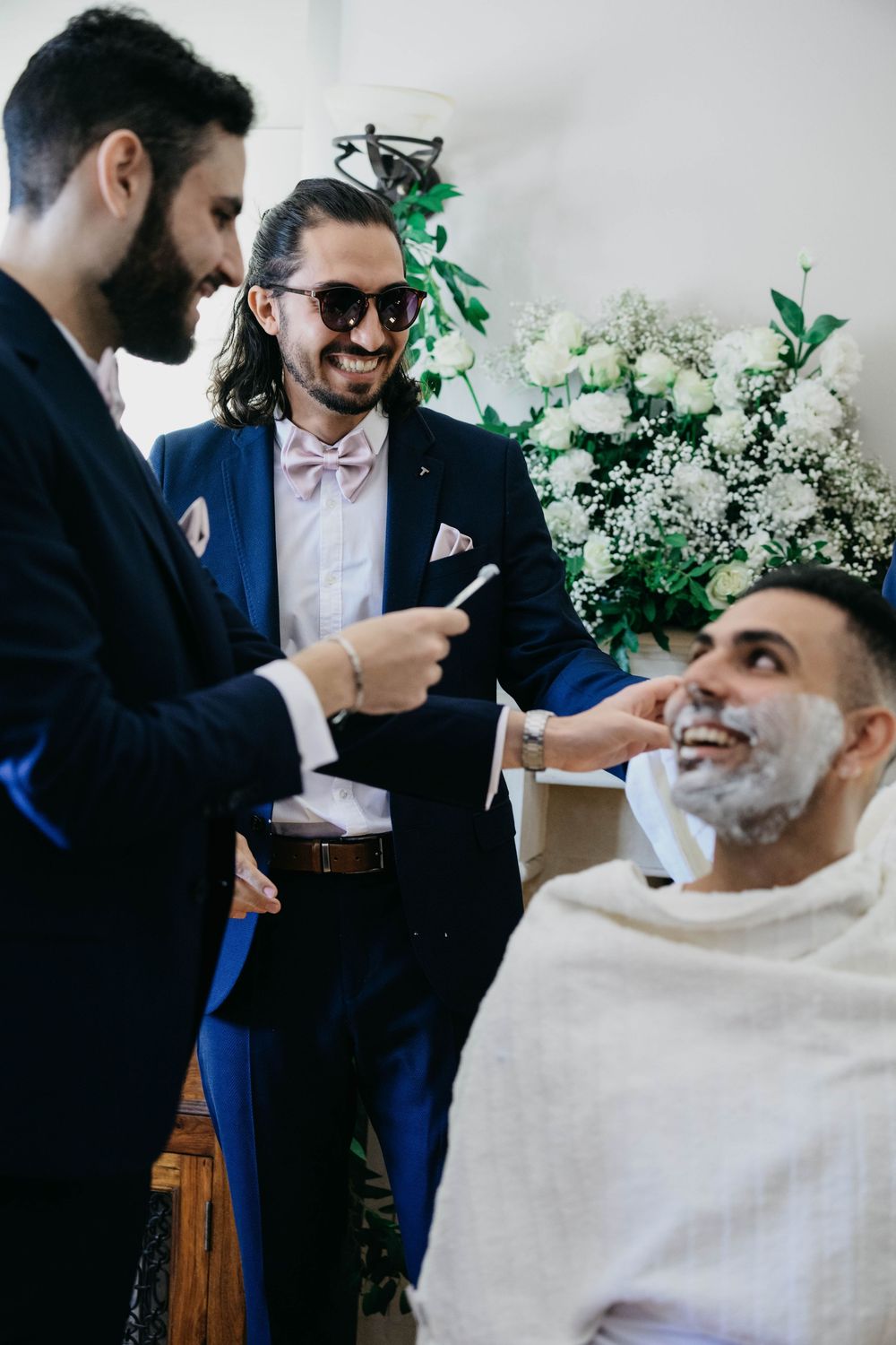 Three people share a candid laughing moment at a wedding preparation scene with fresh white floral arrangements in the background.