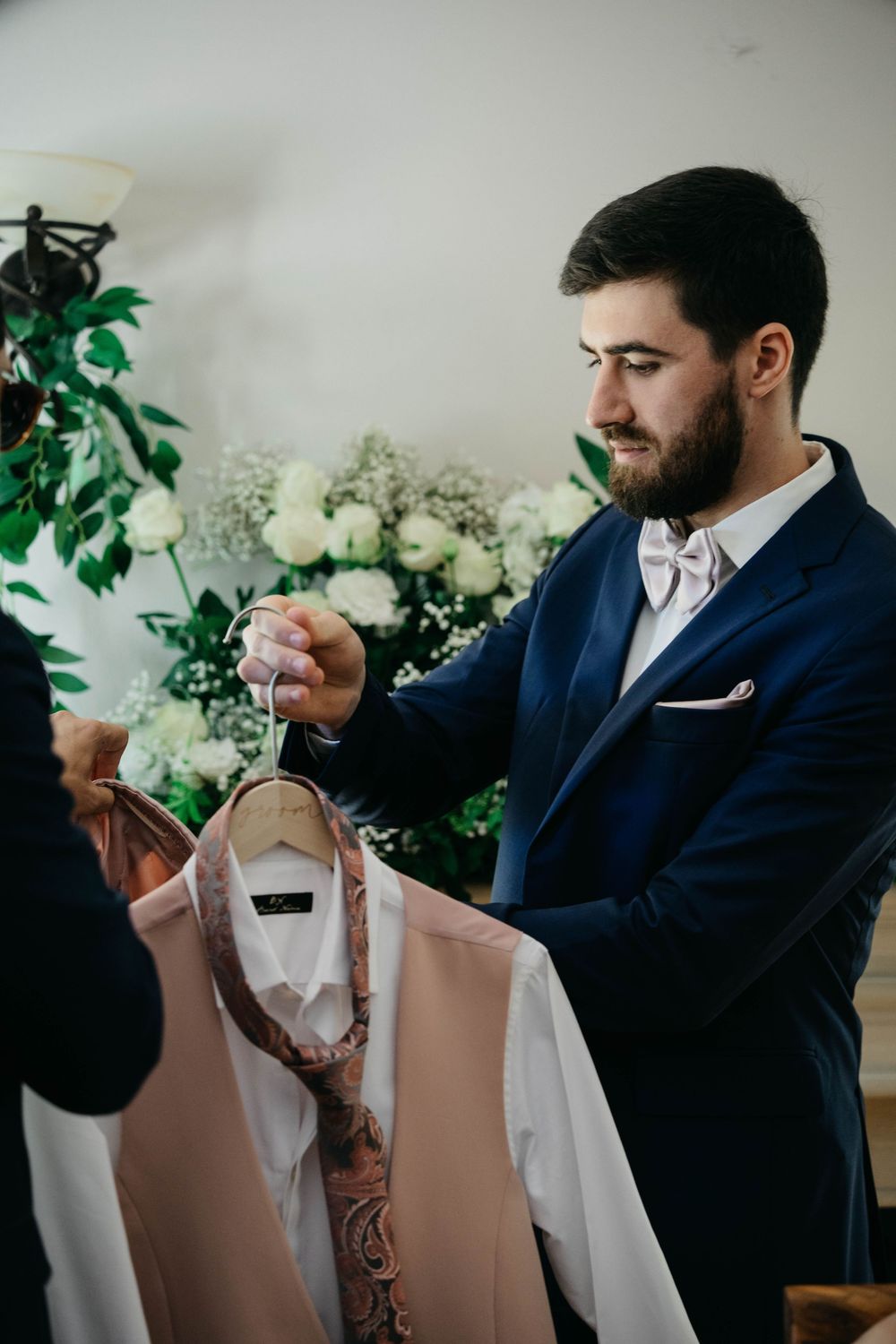 Person in navy suit smells and admires a white wedding gown detail against a light background.