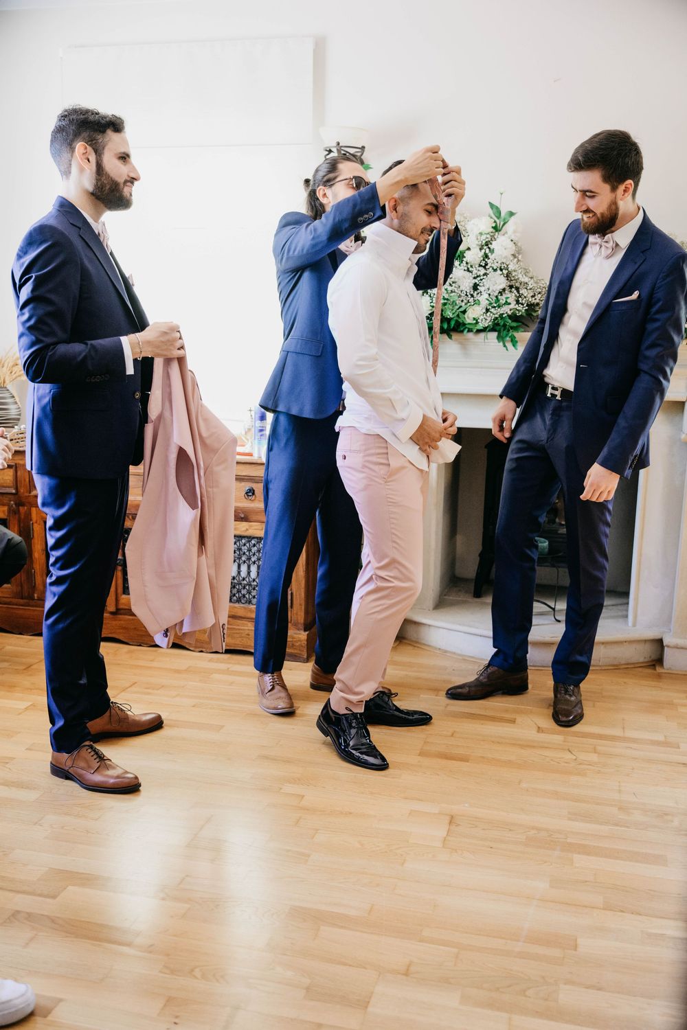 Groomsmen in navy and light pink suits getting ready together in a bright room with hardwood floors.