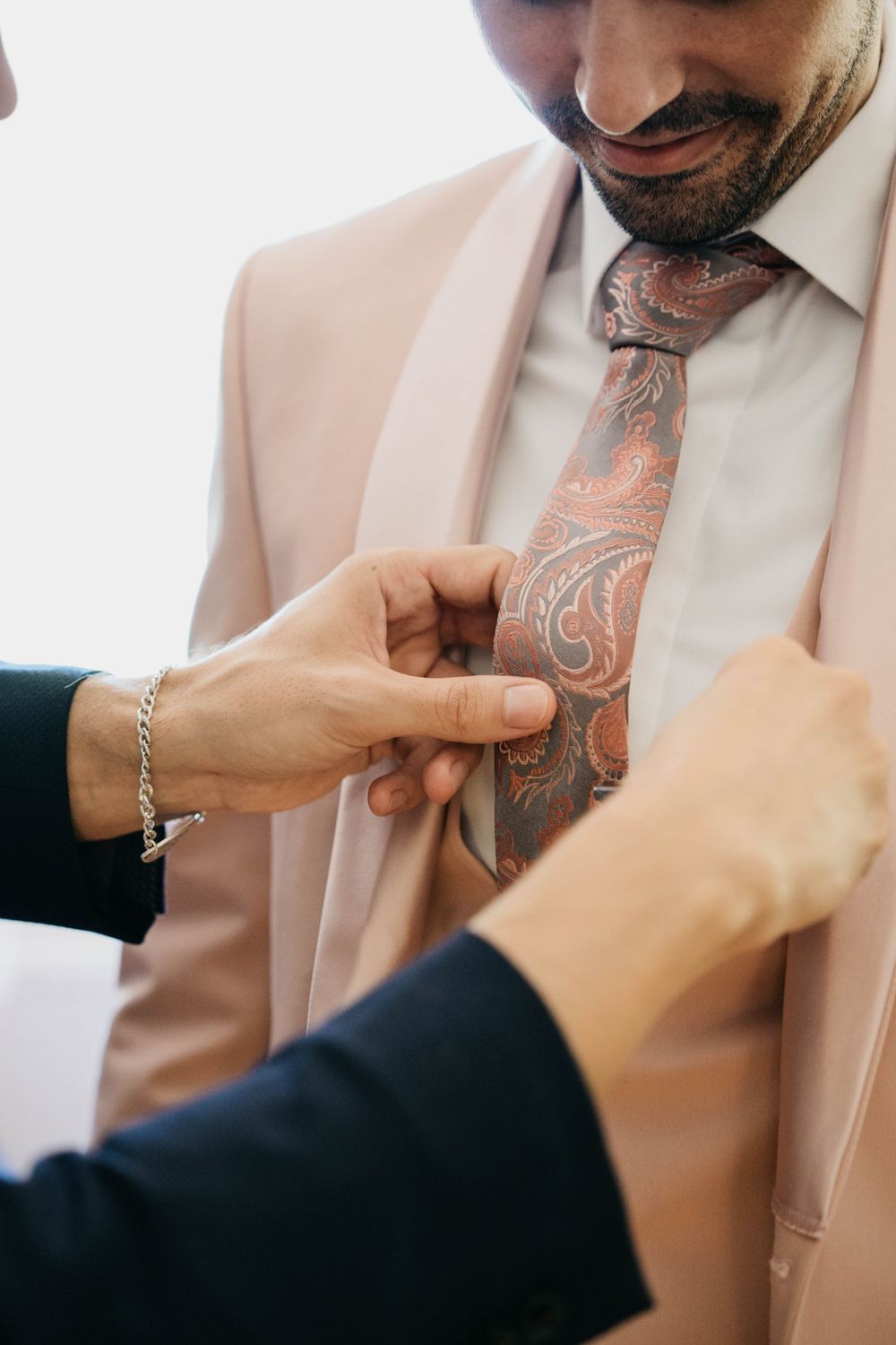 Hands adjusting a pink and silver patterned neck tie against a white dress shirt in a formal setting.