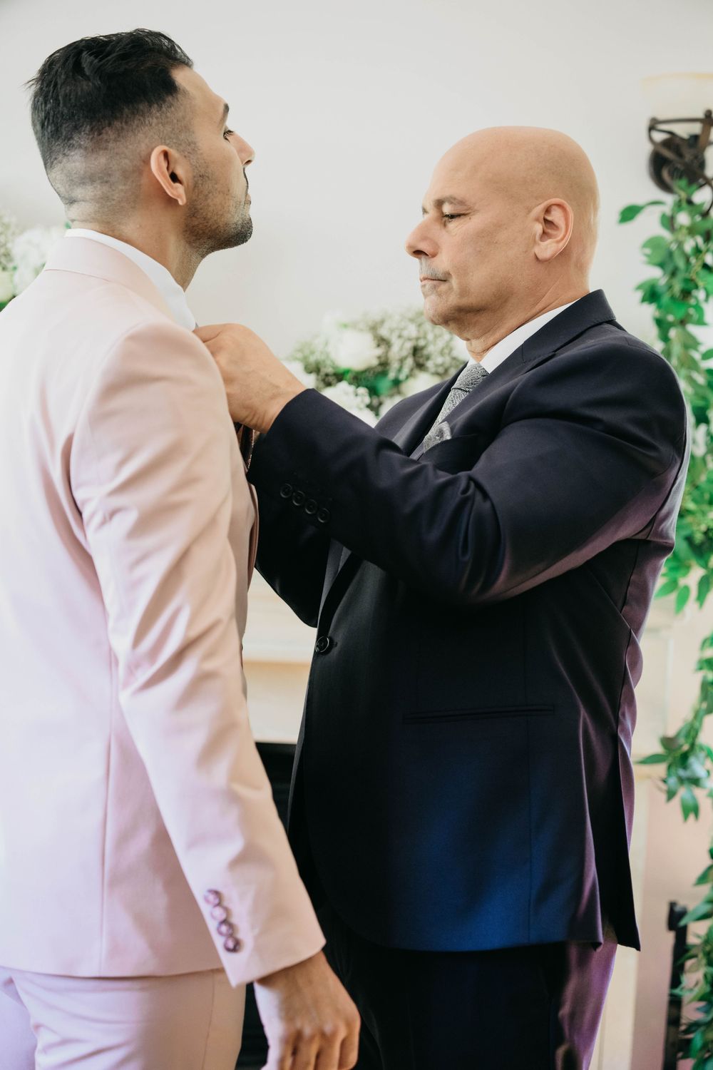 A touching pre-wedding moment as someone helps adjust a bowtie on a person wearing a light pink suit at a wedding.