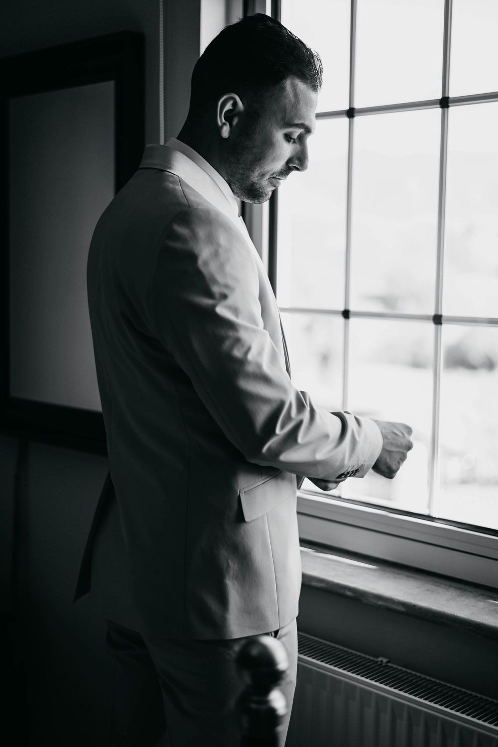 Black and white portrait of a groom in light colored suit gazing thoughtfully out a window on his wedding day.