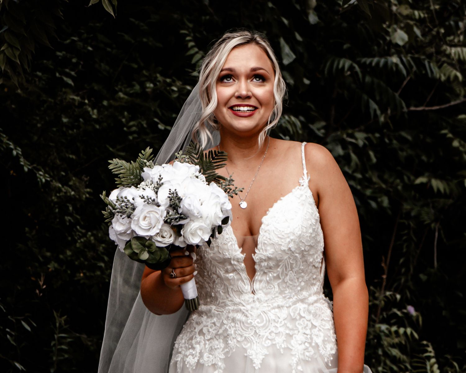 A radiant bride in a white lace dress holds a bouquet of white flowers against a lush garden backdrop.