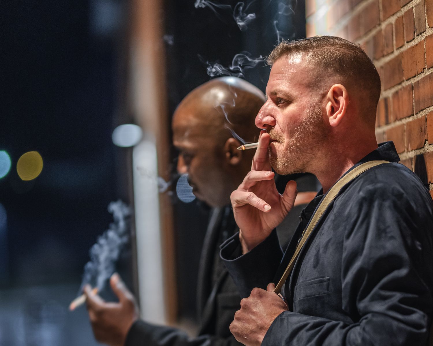 Two people smoking cigarettes against a brick wall at night with bokeh lights in background.