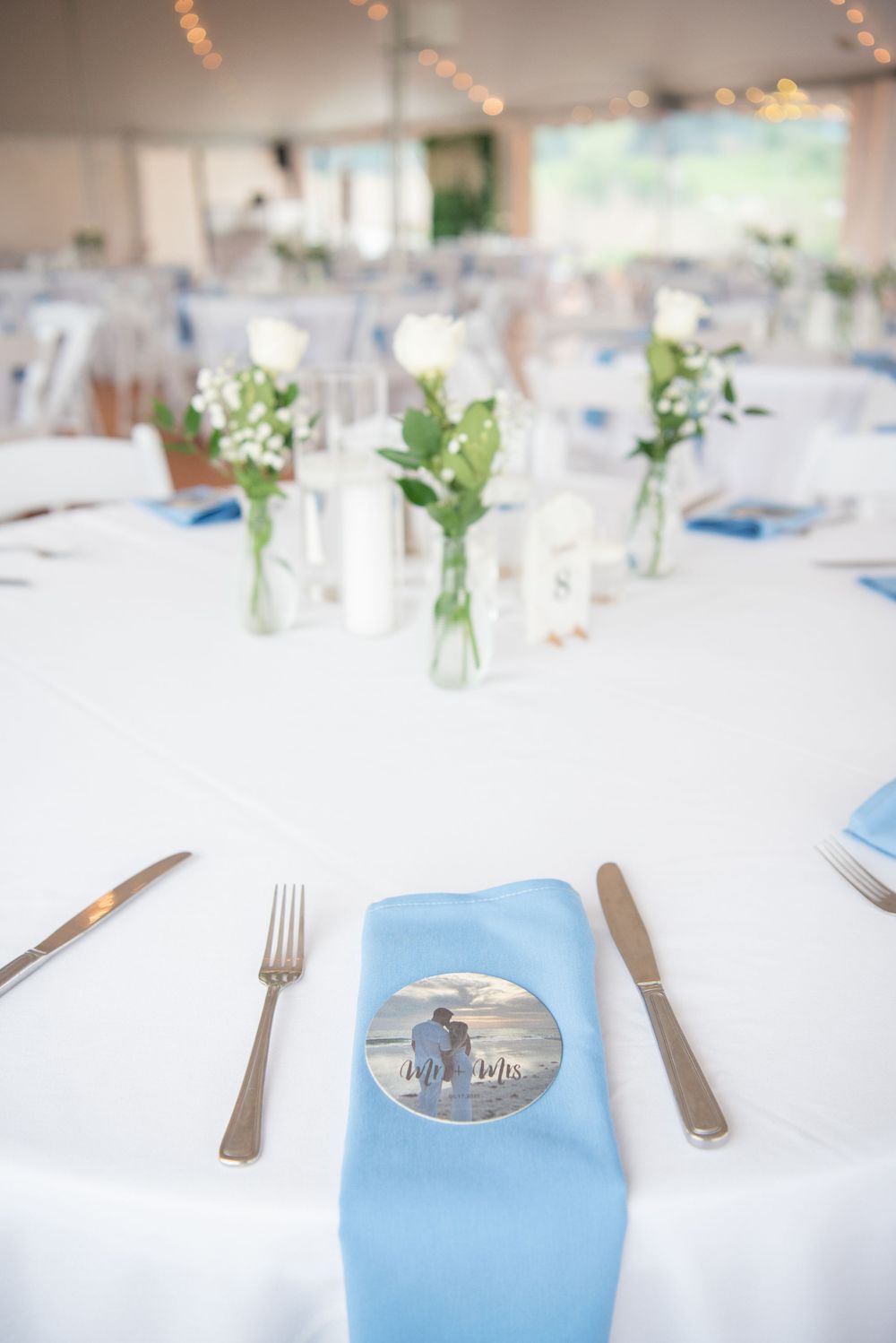 Place settings with silverware and light blue napkins on white tablecloths at a wedding reception.