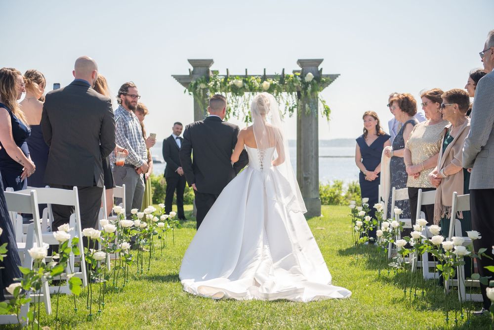 Wedding ceremony guests seated in white chairs on a sunny outdoor lawn with decorative columns.