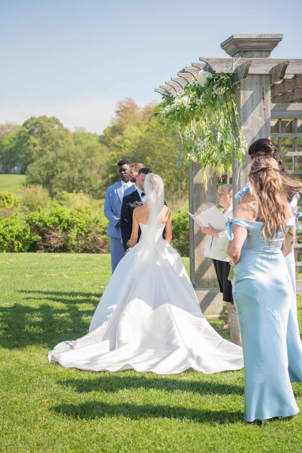 Bridesmaids in light blue dresses attend to the bride's gown on a grassy lawn.