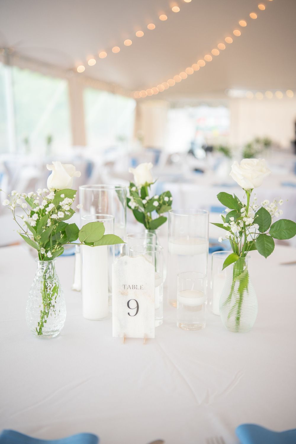 Simple glass bud vases with delicate white flowers line a wedding reception table under soft string lights.