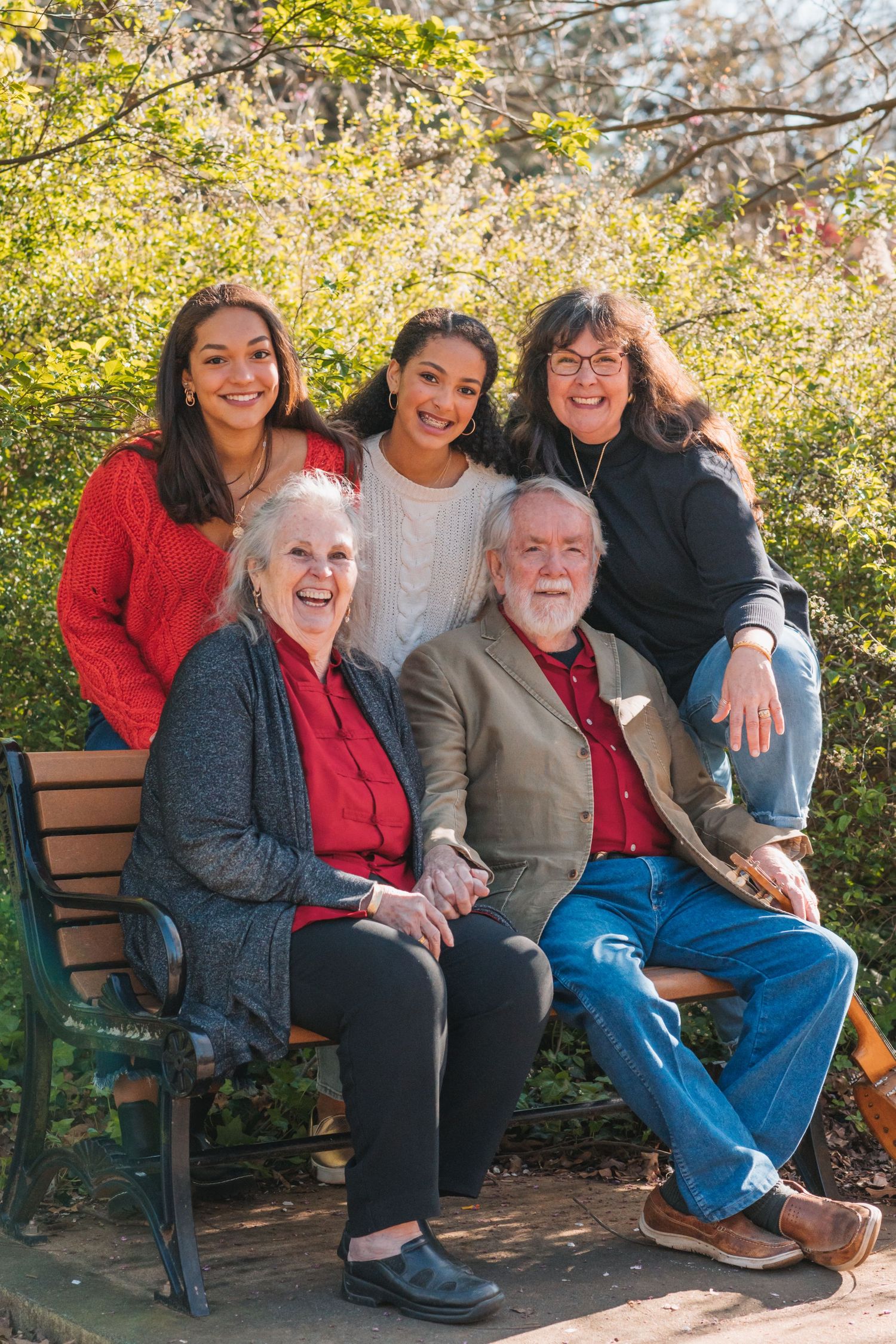 Multigenerational family smiles together while sitting on a bench in a sunny garden setting with blooming trees.
