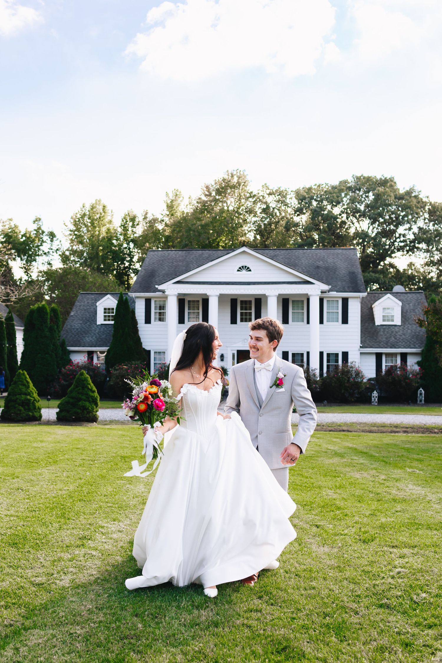 Wedding couple dances and twirls on the lawn in front of a classic white colonial house.