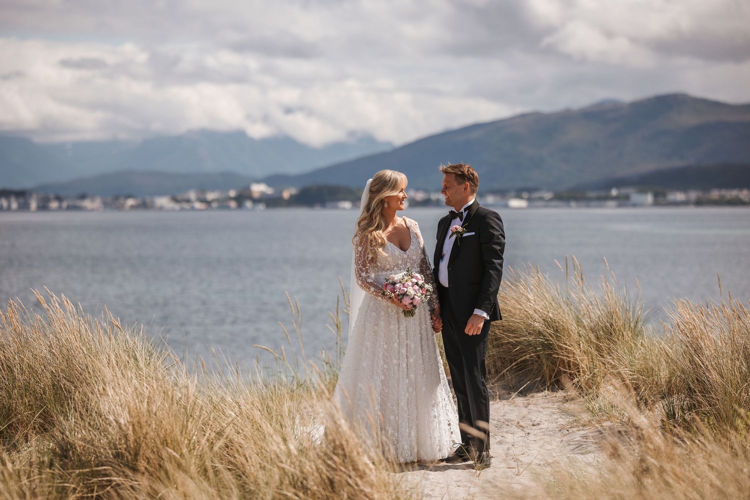 A couple stands together on a coastal path with tall grass and mountains across the water on their wedding day.