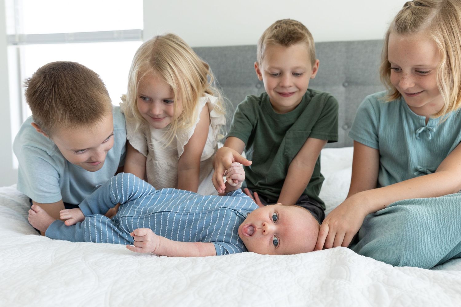 4 kids sitting on a bed looking at their newborn baby boy who is also lying on the bed