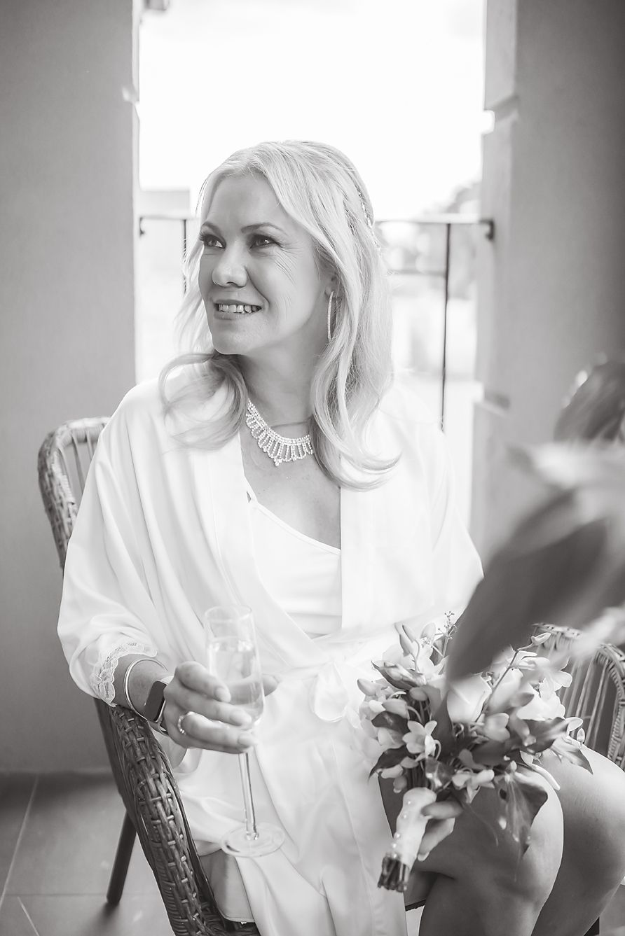 Black and white photo of a bride in white dress and pearl necklace smiling while holding a glass and bouquet.