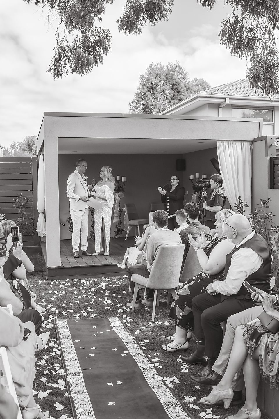 Wedding guests seated outdoors watching ceremony with white flower petals scattered on the ground.