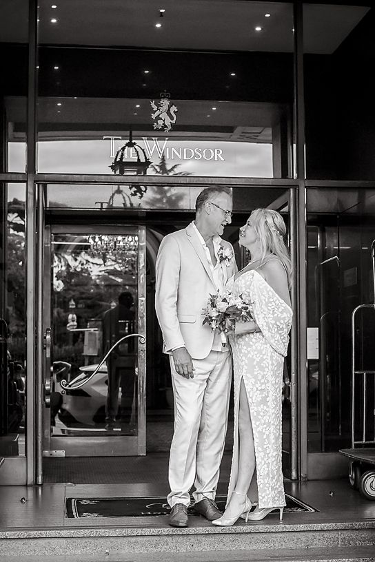 A romantic black and white wedding photo of a couple in matching white outfits standing at the entrance of the Windsor hotel.