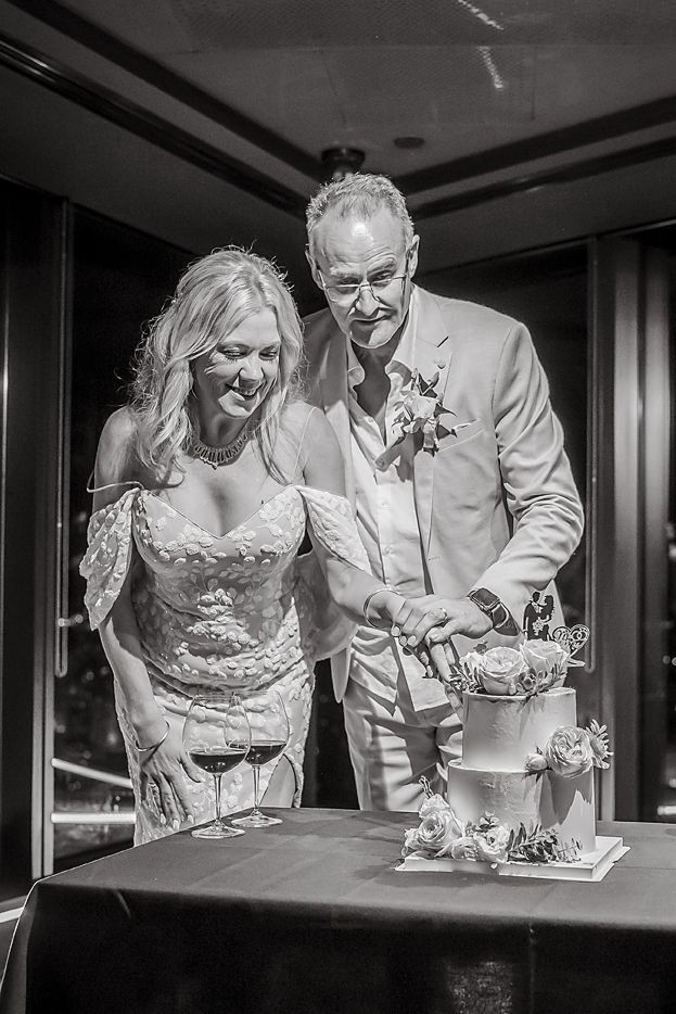 Black and white wedding photo showing a couple slicing their wedding cake together at an elegant reception venue.