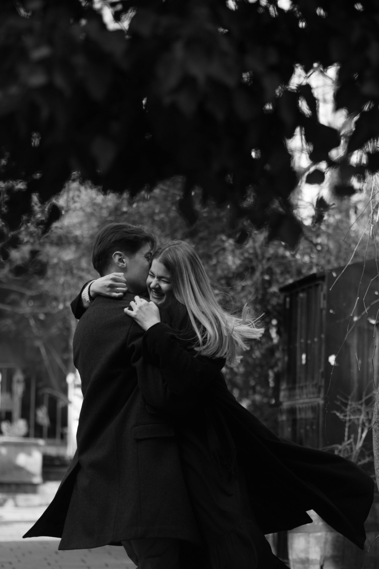 A romantic black and white photograph shows a couple in dark coats embracing and dancing outdoors in a whimsical moment.
