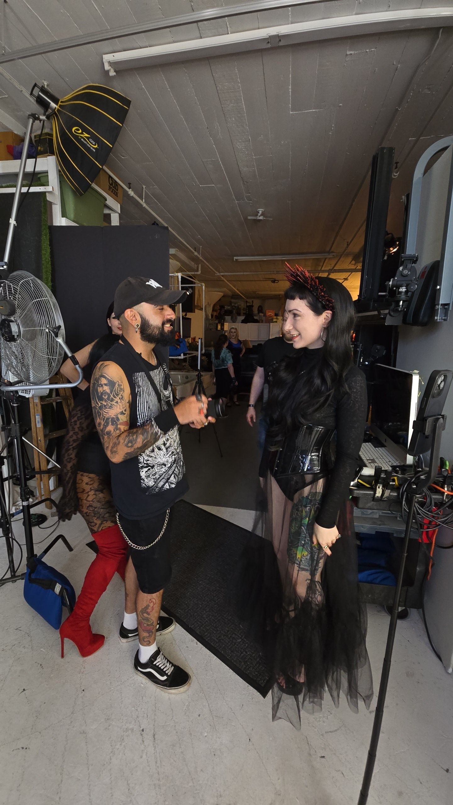 Two people converse in a studio space with photo equipment, one wearing black attire and another in shorts with tattoos.