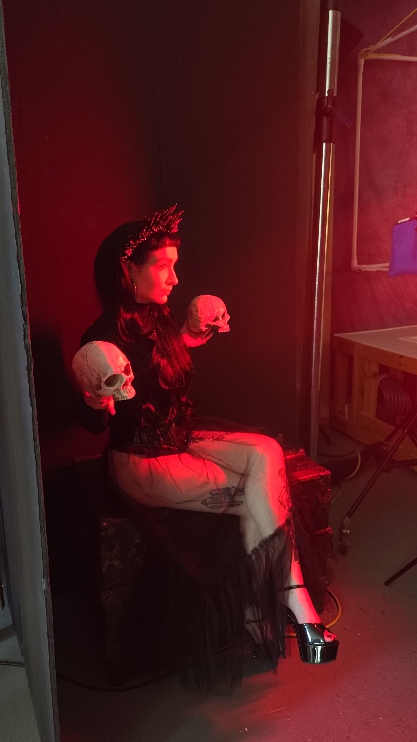 A figure sits in moody red lighting holding two decorative skulls while wearing gothic-style clothing and platform shoes.
