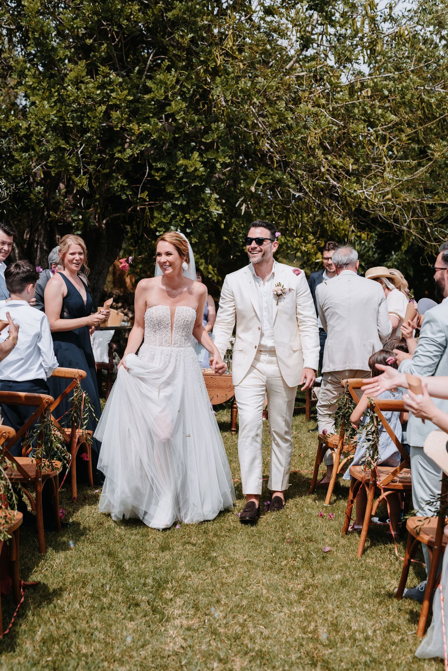 Ein frisch verheiratetes Brautpaar schreitet an einem sonnigen Tag bei einer Mallorcahochzeit zwischen Holzstühlen den Gang hinunter.