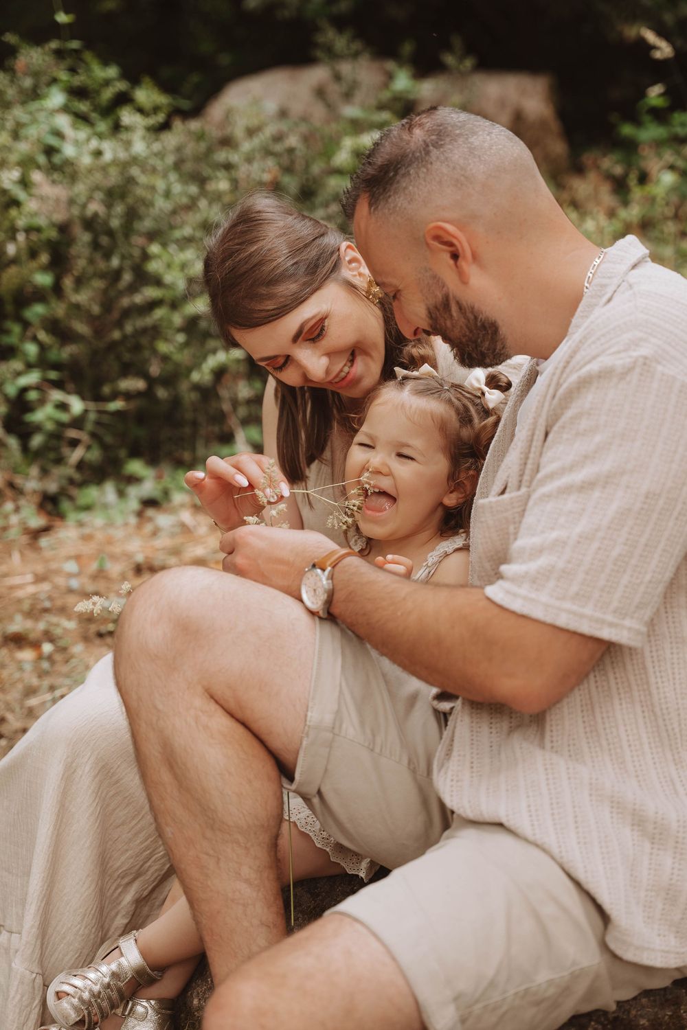 Family shares joyful laughter moment together while sitting outdoors in natural setting wearing neutral colored clothing.