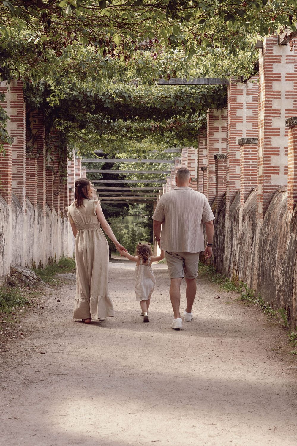 Family walks together holding hands down a scenic garden path lined with brick columns and overhead greenery.