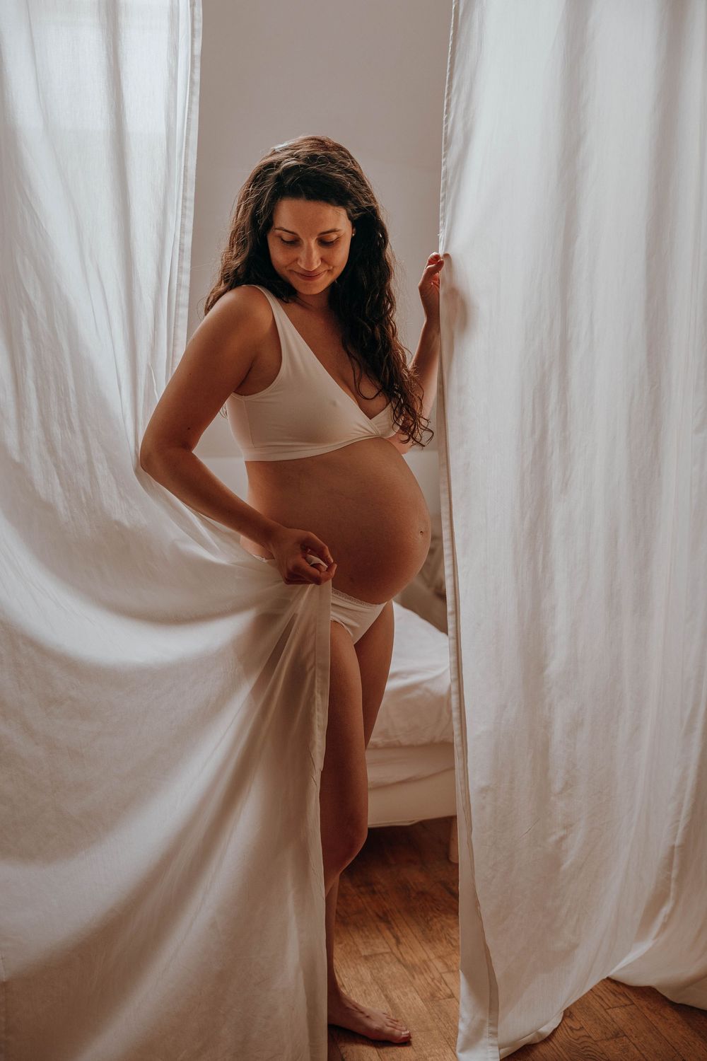Expectant mother in white attire poses between flowing curtains during intimate maternity photoshoot.