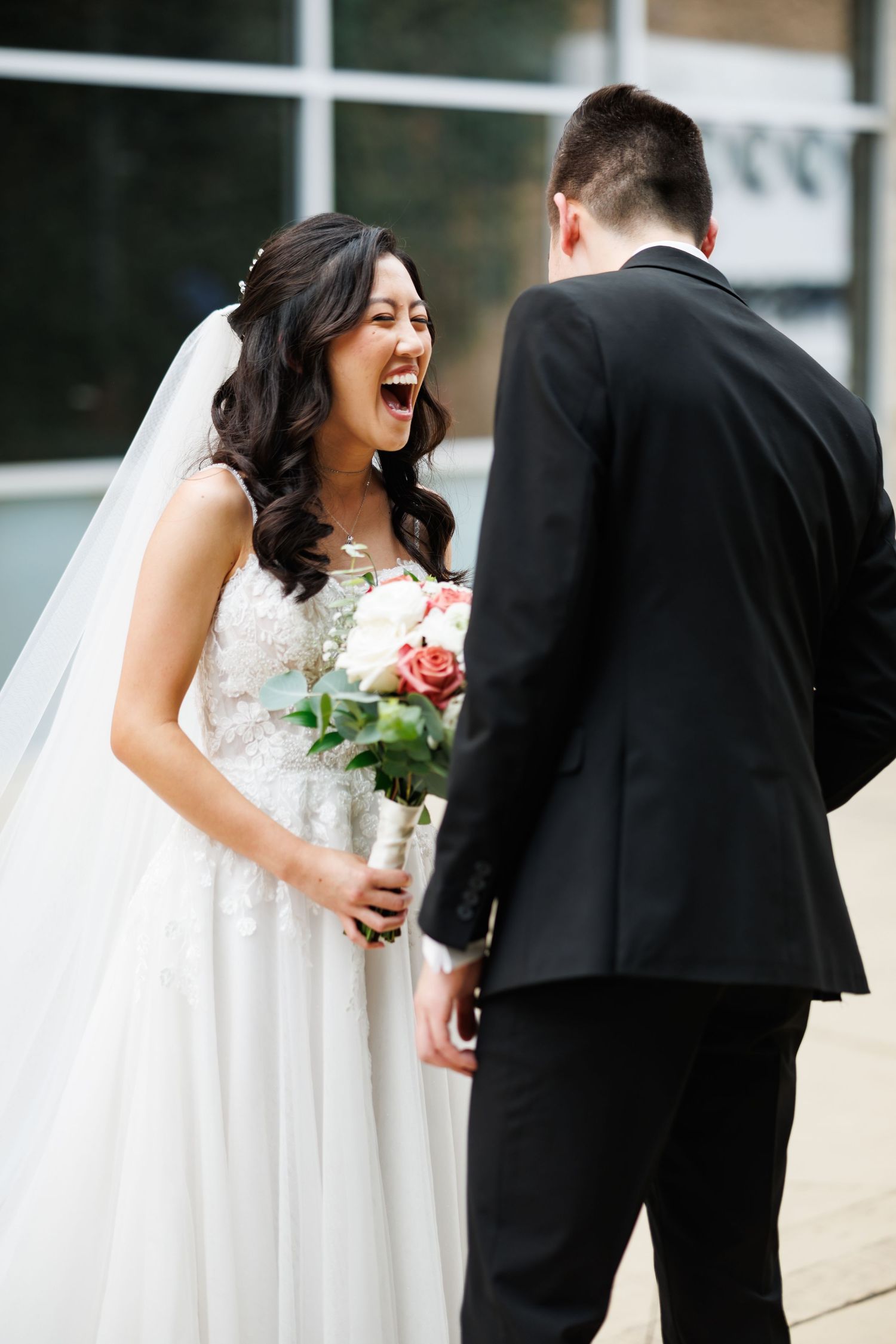 A joyful bride in a white lace gown laughs while holding a bouquet during a wedding first look moment outdoors.