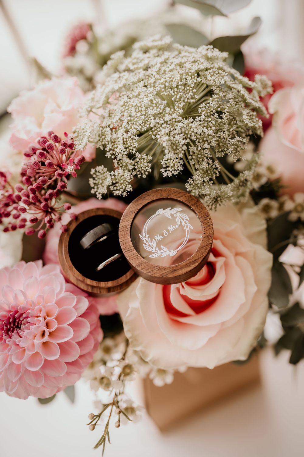 Wedding rings in wooden box nestled among pink roses, dahlias and delicate Queen Anne's lace flowers in a romantic arrangement.